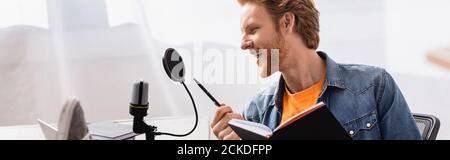 immagine orizzontale dell'emittente eccitata che parla nel microfono mentre si tiene in mano penna e notebook Foto Stock