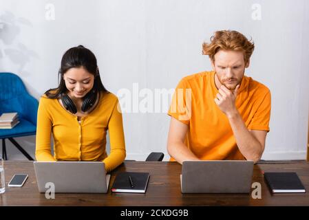 freelance asiatico con cuffie wireless sul collo che utilizzano il laptop vicino uomo pensieroso Foto Stock