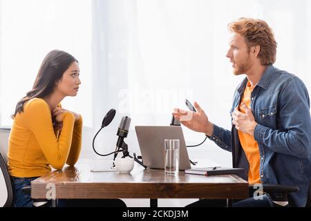 vista laterale dell'intervistatore che gesturing mentre parla con asiatico scioccato donna che tiene le mani vicino al petto Foto Stock