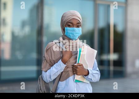 Nero musulmano femmina studente indossare maschera medica in posa all'aperto con cartelle di lavoro Foto Stock