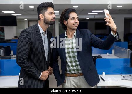Ritratto di due giovani bei imprenditori indiani che guardano al telefono, videoconferenza con il cliente, prendendo selfie, discutendo progetto, Foto Stock
