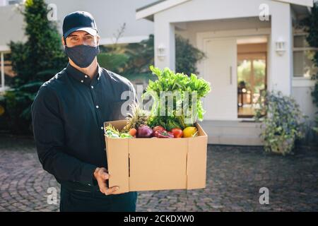 Corrieri maschili che trasportano i generi alimentari ordinati online durante la pandemia. Persona di consegna che indossa una maschera protettiva che fornisce cibo e generi alimentari. Foto Stock