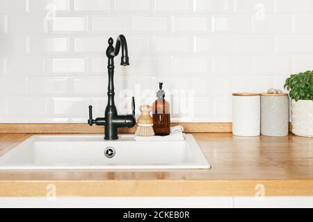 Vista sulla cucina bianca in stile scandinavo, dettagli della cucina, lavello bianco piante di rubinetto nero su tavolo di legno, bianco ceramica muro di mattoni sfondo Foto Stock