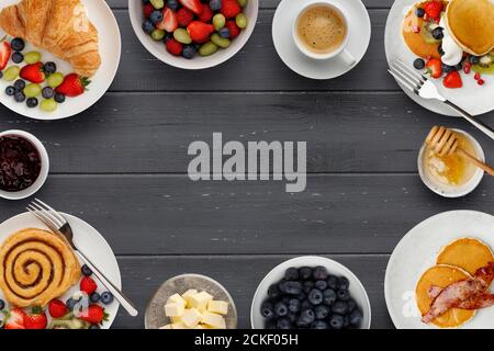 Un bordo di deliziosi piatti per la colazione, pancake e pasterie, ciotole di frutta, marmellata e burro, e una tazza di caffè, su uno sfondo di legno scuro con Foto Stock