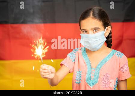 Giovane ragazza Kid sulla maschera medica che tiene Sparkler con tedesco Bandiera come sfondo - concetto che mostra Celebrazione dell'unità tedesca O giorno della Repubblica Foto Stock