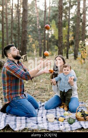 Concetto di famiglia, divertimento e svago. Ritratto di felice famiglia seduta su plaid a scacchi con frutta nella foresta d'autunno. Bel padre, giocolando le mele Foto Stock