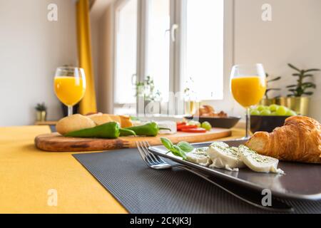 Colazione per due in un moderno e luminoso interno: Baguette fresche a fette su tavola, due peperoni di jalapeno, croissant, mozzarella e succo d'arancia Foto Stock