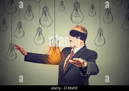 Uomo anziano bendato che cammina attraverso le lampadine alla ricerca di luce brillante idea isolata su sfondo grigio Foto Stock