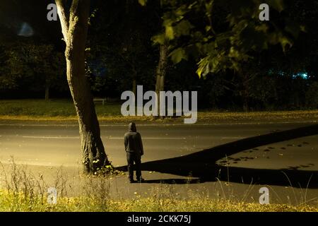 Guarda giù su una misteriosa figura incappucciata in piedi su una strada. Sotto un albero di notte Foto Stock