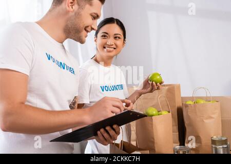 Fuoco selettivo del volontario asiatico che tiene la mela vicino all'uomo con appunti e pacchetti nel centro di beneficenza Foto Stock