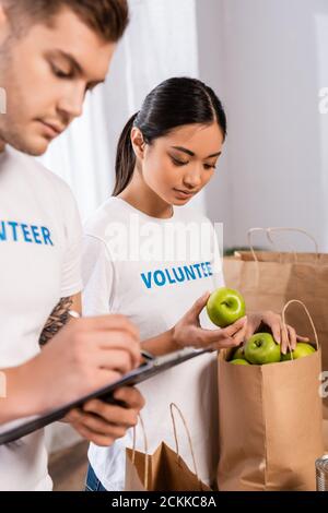 Fuoco selettivo del volontario asiatico che tiene le mele vicino alla scrittura dell'uomo sugli appunti nel centro di beneficenza Foto Stock