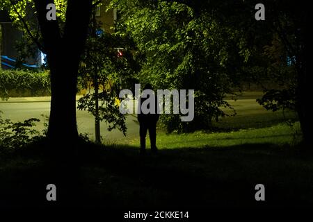 Guardando giù su una misteriosa figura incappucciata in piedi su una strada. Sotto un albero di notte Foto Stock