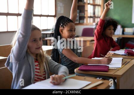 Gruppo di bambini che alzano le mani mentre si siedono sul loro scrivania a scuola Foto Stock