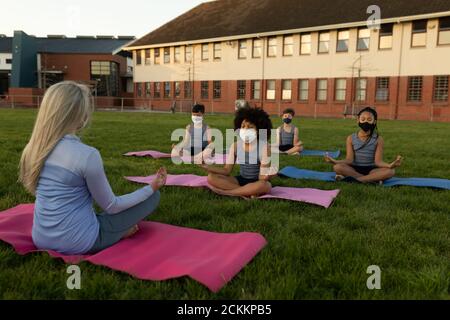 Insegnante femminile e gruppo di bambini che indossano maschera che si esibiscono yoga in giardino Foto Stock