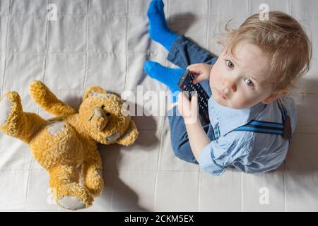 Cute baby boy e il suo orsacchiotto che guarda la TV seduto su un divano nel soggiorno di casa, vista dall'alto Foto Stock