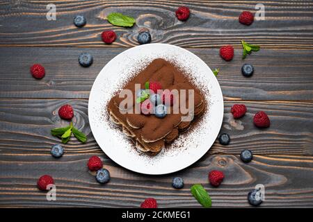 Porzione di tipico dessert tiramisù con lamponi e mirtilli sfondo di legno Foto Stock