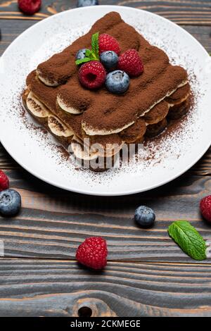 Porzione di tipico dessert tiramisù con lamponi e mirtilli sfondo di legno Foto Stock