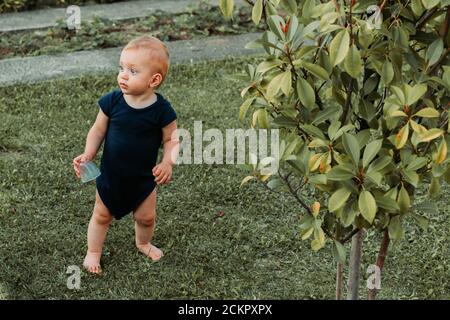 Il bambino piccolo in bodysuit si trova a piedi nudi nel giardino guardando qualcosa Foto Stock