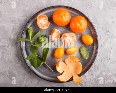 tangerini, kumquat e un ramo di menta su una piastra di ceramica su uno sfondo grigio in cemento, vista dall'alto, posa piatta, primo piano. Foto Stock