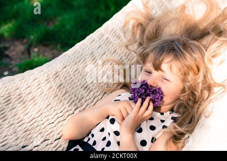 bella ragazza bionda con lunghi capelli biondi giacenti in bianco amaca e lilla odore fiore Foto Stock