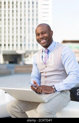 Elegante uomo d'affari nero sorridente. È seduto e ha un computer portatile sulle gambe. Concetto aziendale. Foto Stock