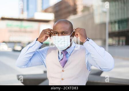 Ritratto di un uomo nero che mette sul viso una maschera medica. È all'aperto in città. Concetto di nuova normalità. Foto Stock