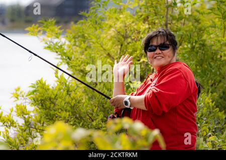 Una donna asiatica di mezza età sorridente, negli anni '50, tiene una canna da pesca e fa un segno V. Cespugli verdi sullo sfondo Foto Stock