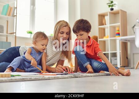 La mamma e i bambini giovani disegnano insieme un quadro seduto sul pavimento in asilo nido Foto Stock