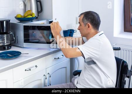 Giovane uomo in Whellchair che usa forno a microonde per cuocere in cucina. Mettere a fuoco sulla sua mano. Foto Stock