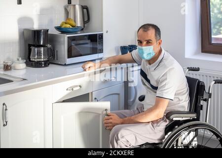Sorridente giovane uomo portatore seduto su sedia a rotelle in cucina. Giovane uomo che indossa la maschera seduta di fronte alla cucina. Mettere a fuoco sul suo volto. Foto Stock