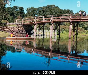 Una canoa contenente tre turisti passa sotto lo storico North Bridge di Concord, Massachusetts. Sopra c'è un cielo blu profondo, sotto il verde e il blu profondo Foto Stock