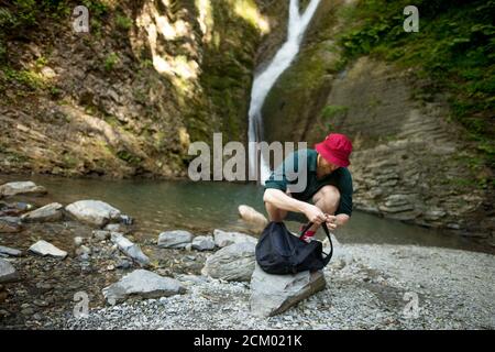 Uomo turistico in cappello rosso e abbigliamento estivo toglie qualcosa dal suo zaino vicino alla bella natura e cascata sfondo Foto Stock