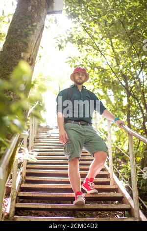 Giovane uomo barbuto in Red Hat sorge su scale di legno nel bosco selvatico. Basso angolo vista del viaggiatore di tela che indossa pantaloni corti e t-shirt, e rosso sneakers. Foto Stock