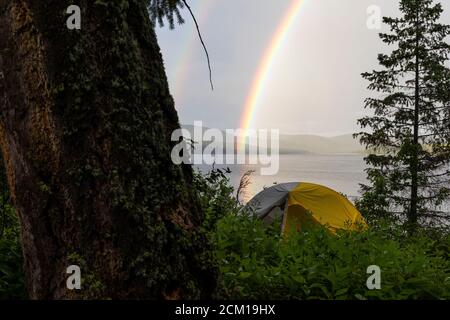 Tenda in cespugli vicino alberi sulla riva con arcobaleno vivido sul lago calmo Foto Stock
