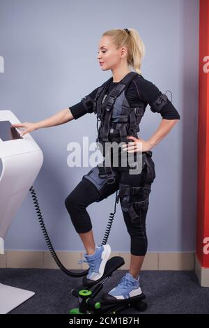 donna in grande vestibilità che indossa una tuta ems su stepper e regola l'allenamento nel centro sportivo. concetto di salute e tempo libero. foto a lunghezza intera Foto Stock