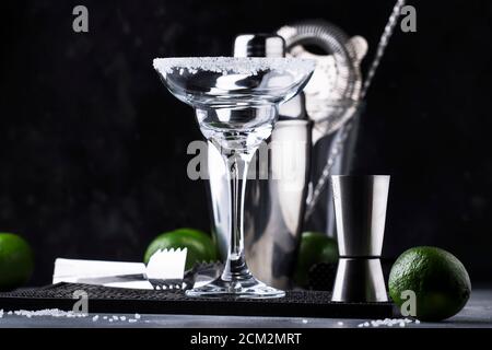 Concetto di preparazione dei cocktail. Svuotare il bicchiere da cocktail margarita, lo shaker e gli utensili da bar, il lime e il sale sul banco bar scuro Foto Stock