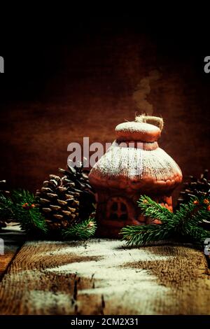 Casa di Natale di argilla con coni di pino e rami di abete sotto la neve, sfondo di legno d'annata, fuoco selettivo Foto Stock