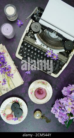 Spazio di lavoro desktop per scrittori vintage a tema viola con macchina da scrivere, tè lavendar, fiori di hibiscus e libri antichi su elegante sfondo viola testurizzato. Da a. Foto Stock