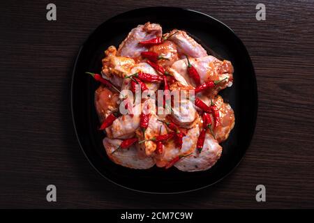 Ali di pollo crudo in una marinata con spezie su un piatto nero, un sacco di peperone rosso caldo, su un tavolo di legno scuro, vista dall'alto, concetto di cibo Foto Stock