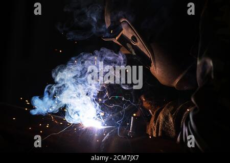 Saldatore al lavoro. Uomo in una maschera protettiva. La saldatrice realizza giunzioni sul metallo. Scintille e fumo durante la saldatura. Foto Stock