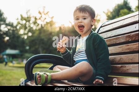 Ragazzo caucasico seduto in panchina nel parco sta mangiando un gelato e sorridi alla macchina fotografica con la bocca sporca Foto Stock