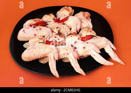 Ali di pollo crudo con spezie. Pollo crudo sottaceto con pepe caldo, su piatto nero, fondo luminoso Foto Stock