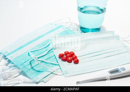 Maschera medica per prevenire la diffusione del virus COVID-19, una manciata di compresse rosse, un termometro e un bicchiere d'acqua. Protezione antivirale monouso Foto Stock