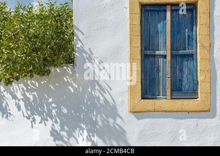 La finestra di una casa greca su un muro bianco Con finiture gialle e persiane blu in legno su un Sunny giorno Foto Stock