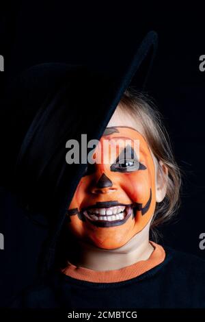 Bambino sorridente con un cappello nero e un motivo di zucca sulla sua faccia su uno sfondo nero, Halloween Foto Stock