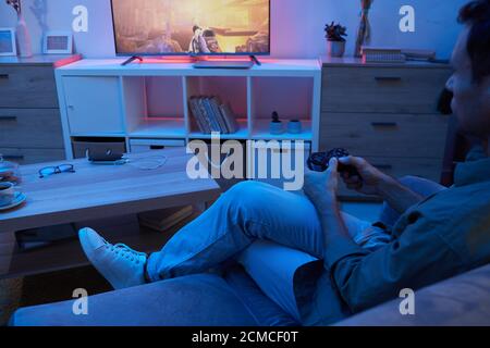 Vista posteriore del giovane seduto sul divano e giocando in videogioco in camera buia a casa Foto Stock
