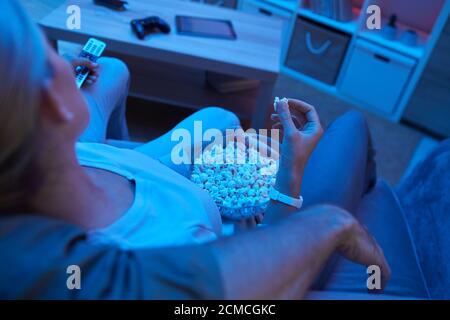Vista posteriore della giovane donna che mangia popcorn mentre il suo ragazzo abbracciandola sul divano che guardano un film insieme Foto Stock