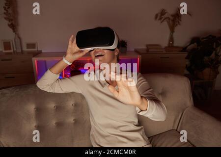 Giovane donna seduta sul divano in occhiali virtuali che giocano gioco virtuale a casa Foto Stock