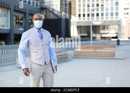 Uomo nero intraprendente sulla strada. Indossa una maschera facciale e tiene un laptop sotto il braccio. Concetto di nuova normalità nel contesto della pandemia di coronavirus. Foto Stock