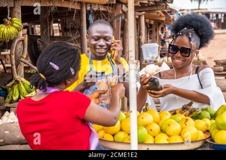 giovane uomo nero che fa una telefonata e tiene una carta di credito, donna che paga per cibo roba in un mercato africano locale Foto Stock